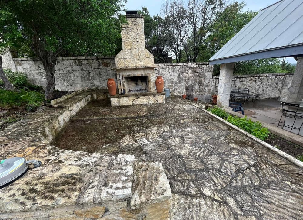 Outdoor patio with stone fireplace, planters, and seating area surrounded by greenery.